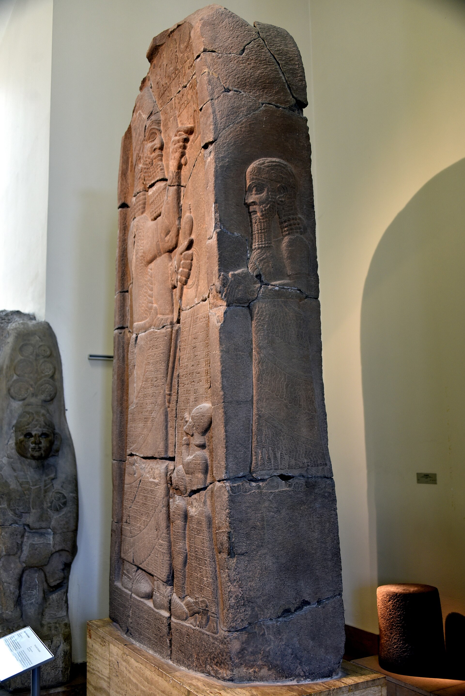 This commemorative basalt stela depicts the Assyrian king Esarhaddon worshiping gods and symbols of gods. The king's left hand holds a royal mace and two ropes. These ropes pass through the lips of two captives. The knee