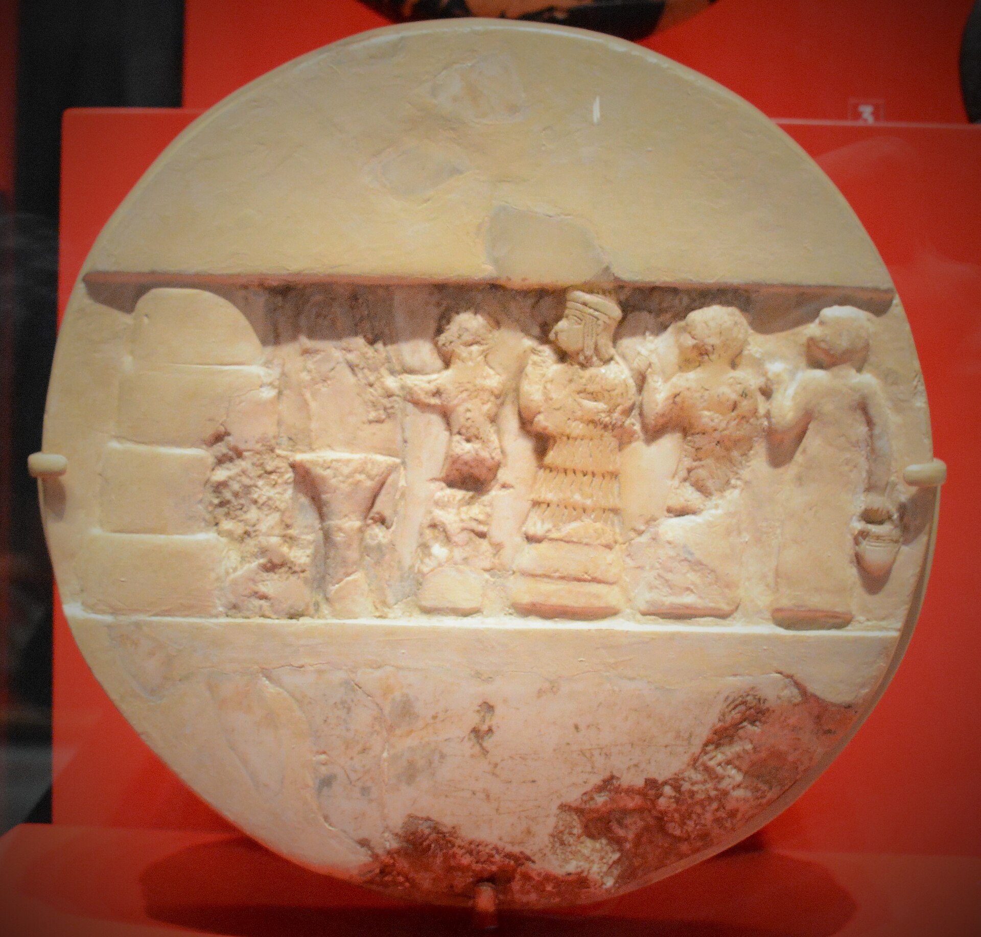Disque d'Enheduanna. Albâtre, v. 2350-2300 av. J.-C. Ur. Diamètre : 25.6 cm, épaisseur : 7.1 cm. Description du bas-relief : scène rituelle, un prêtre fait une libation devant un autel à quatre étages (à gauche), accompa