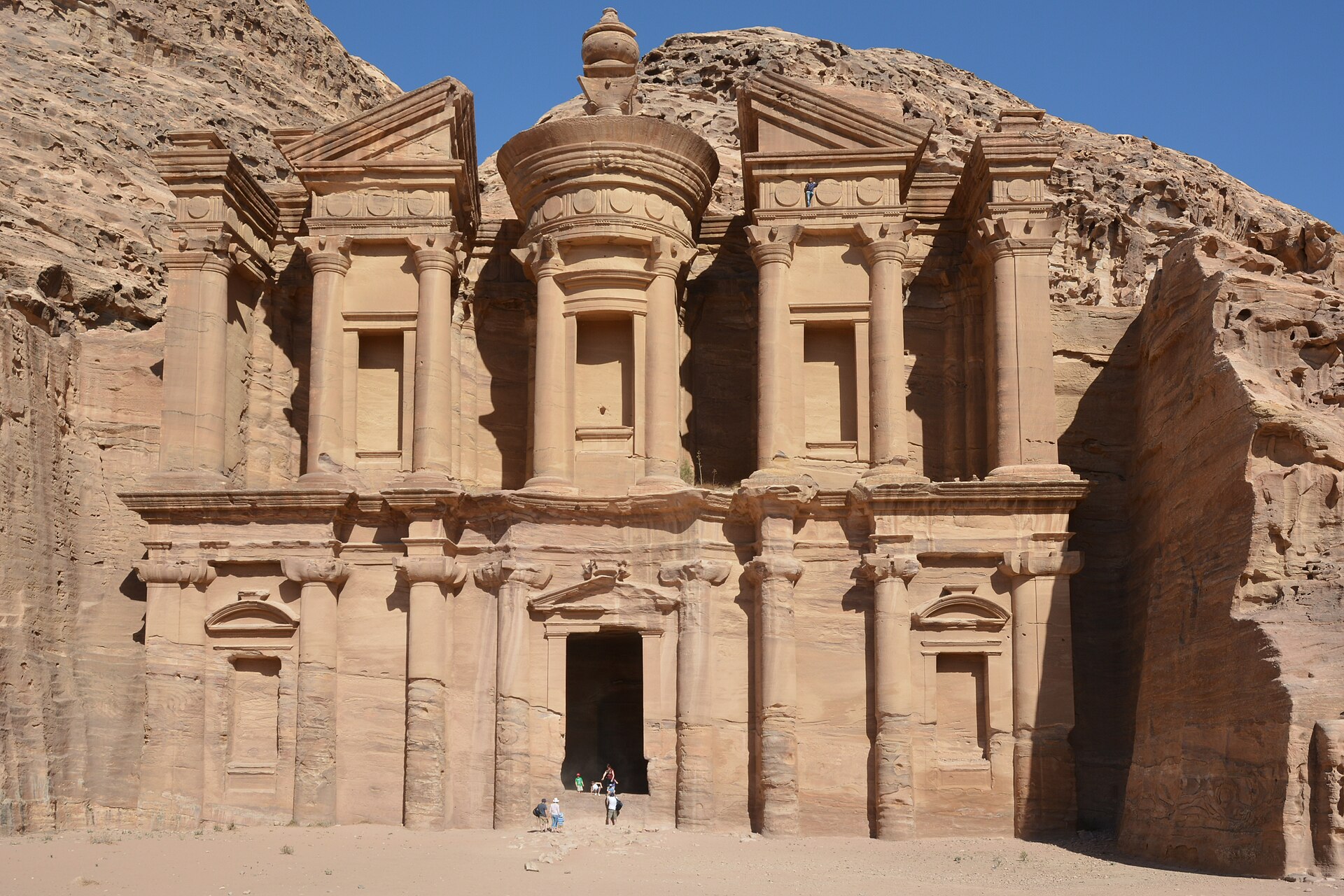 Al Deir, "The Monastery", a monument closely resembling Al Khazneh though less ornate. It can be accessed climbing a long flight of stairs carved in the mountain above Petra. A man can be seen at the top of the facade, s